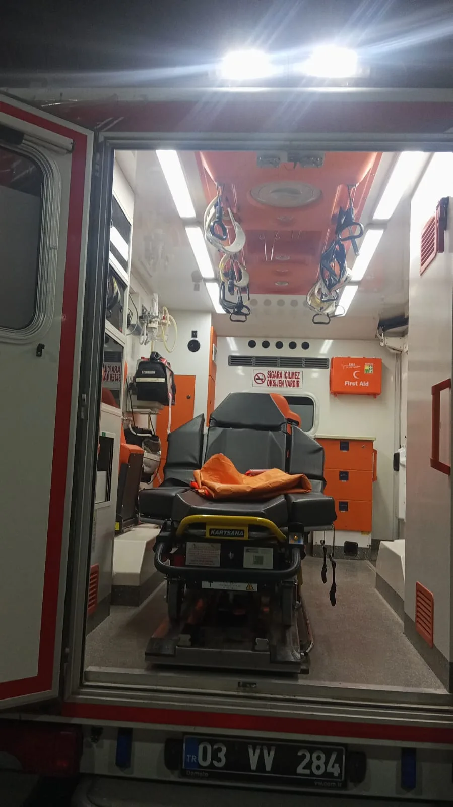 Obez/bariyatrik ambulansın hasta bölmesi: KARTSANA BRAVA elektronik sedye, tavan tutamakları, ilk yardım dolabı ve turuncu saklama dolabı — birimin kendi aracından, yüz içermez